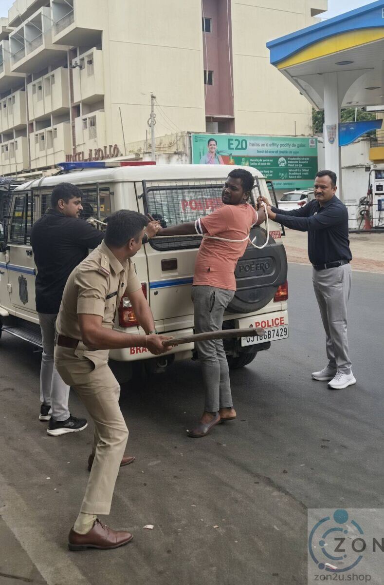 A man tied with rope to the back of a police vehicle while an officer in uniform holds a wooden lathi